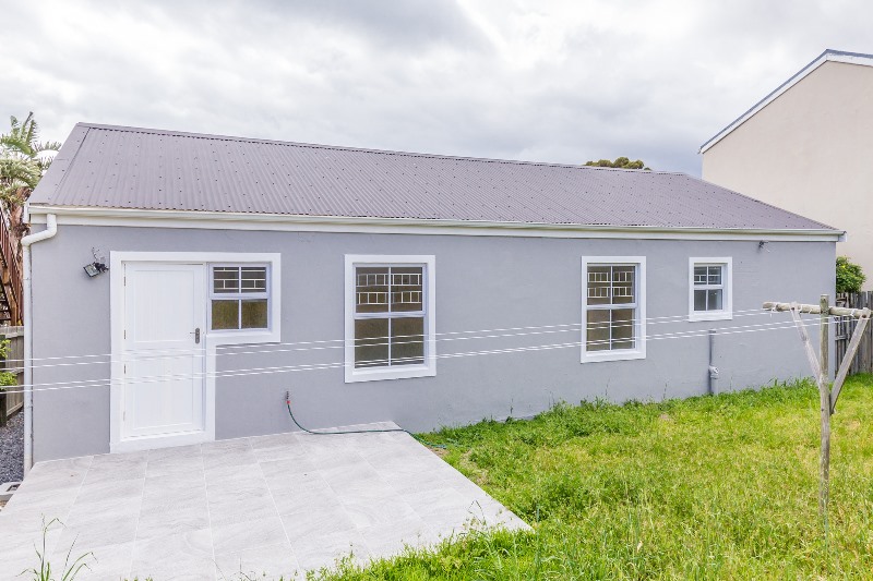 The rear of the house with a dark metal roof and a bare patio slab in an overgrown yard